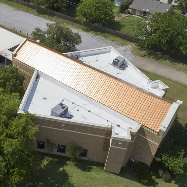 commercial-hero - Linear Roofing - A Stronghouse Partner Aerial view of a brick building with a long orange metal roof running along its top, several HVAC units on the roof, and surrounding trees and a street.