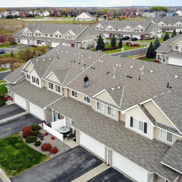 multi-family-hero - Linear Roofing - A Stronghouse Partner Aerial view of a townhouse community with beige siding and gray shingle roofs; a worker stands on a roof inspecting the shingles.