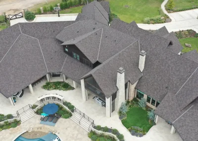 residential-roofing-01 - Linear Roofing - A Stronghouse Partner Aerial view of a large house with a dark multi-gabled roof, stone chimneys, and glass entry, surrounded by a curved patio, landscaped lawns, and a curved blue swimming pool with lounge chairs and an umbrella.