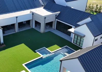 residential-roofing-05 - Linear Roofing - A Stronghouse Partner Aerial view of a modern white house with blue metal roofs surrounding a rectangular pool and green lawn, with lounge chairs nearby.