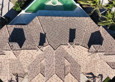 residential-roofing-06 - Linear Roofing - A Stronghouse Partner Aerial view of a large house with tan-brown tiled roof and multiple pitched sections, with a kidney-shaped green pool at the top edge and palm trees nearby.