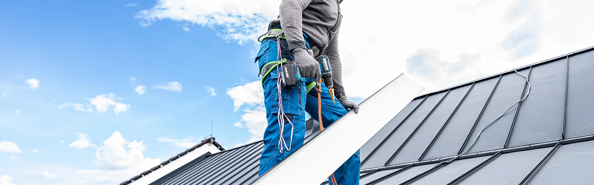 A construction worker on a sloped metal roof, securing a white panel with a drill and tools at his belt.