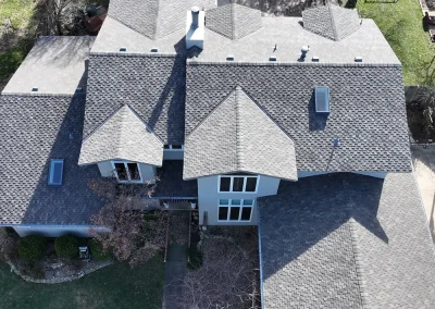storm-restoration-01 - Linear Roofing - A Stronghouse Partner Aerial view of a large gray-shingled house with multiple intersecting roofs, chimneys, and skylights, surrounded by a yard and trees.
